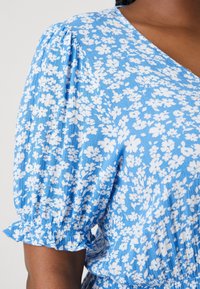 Vestido floral azul con escote en V, mangas abullonadas y puños acampanados. La tela presenta un patrón floral blanco sobre un fondo azul.