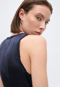 Γυναίκα με κοντά καστανά μαλλιά, φορώντας χρυσό σκουλαρίκι κρίκο και αμάνικο σκούρο ναυτικό τοπ, ρίχνοντας μια ματιά πίσω από τον ώμο της πάνω σε απλό φόντο.