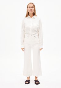 Chemise blanche à manches longues, pantalon large taille haute blanc, et sandales noires à enfiler. Design minimaliste avec texture lisse.
