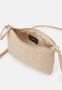 Borsa a tracolla beige trapuntata con chiusura a zip e un cinturino per il polso. Texture morbida con motivi a onda. L'interno include un'etichetta con logo.