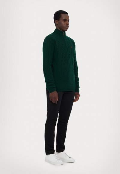 Green cable-knit sweater with high collar, featuring a zip closure and small logo on the chest, paired with black pants and white sneakers.