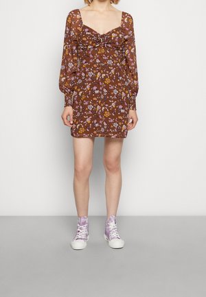 Frau trägt ein braunes, langärmeliges Minikleid mit Blumenmuster und aufgepolsterten Ärmeln sowie lila High-Top-Sneaker, steht vor einem einfarbigen Hintergrund.