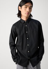 Schwarze button-up Hemd mit spitzem Kragen und langen Ärmeln, ausgestattet mit kontrastierenden weißen Knöpfen und einem kleinen Logo auf der Brust.
