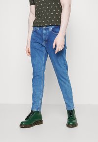 Ljusblå jeans med slim fit, som har klassiska designdetaljer, kombinerat med mörkgröna snörstövlar och en mönstrad mörkgrön skjorta.