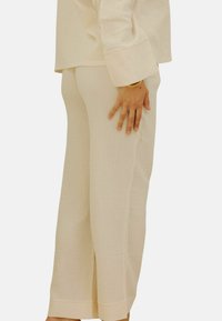Pantalons en coton texturé de couleur beige clair, avec une coupe large, des fentes sur les côtés à l'ourlet et une coupe décontractée. Une main repose sur la hanche pour référence.