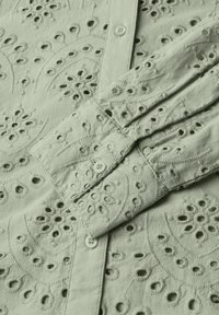Camicia ricamata di colore verde chiaro con motivi floreali, dettagli a intaglio e chiusura con bottoni. Stoffa morbida con accenti testurizzati.