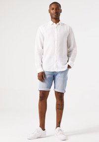 Witte linnen overhemd met knopen, lichtblauwe opgeknoopte denim short, witte sneakers en zichtbare tattoo op het onderbeen. Eenvoudige hedendaagse stijl.