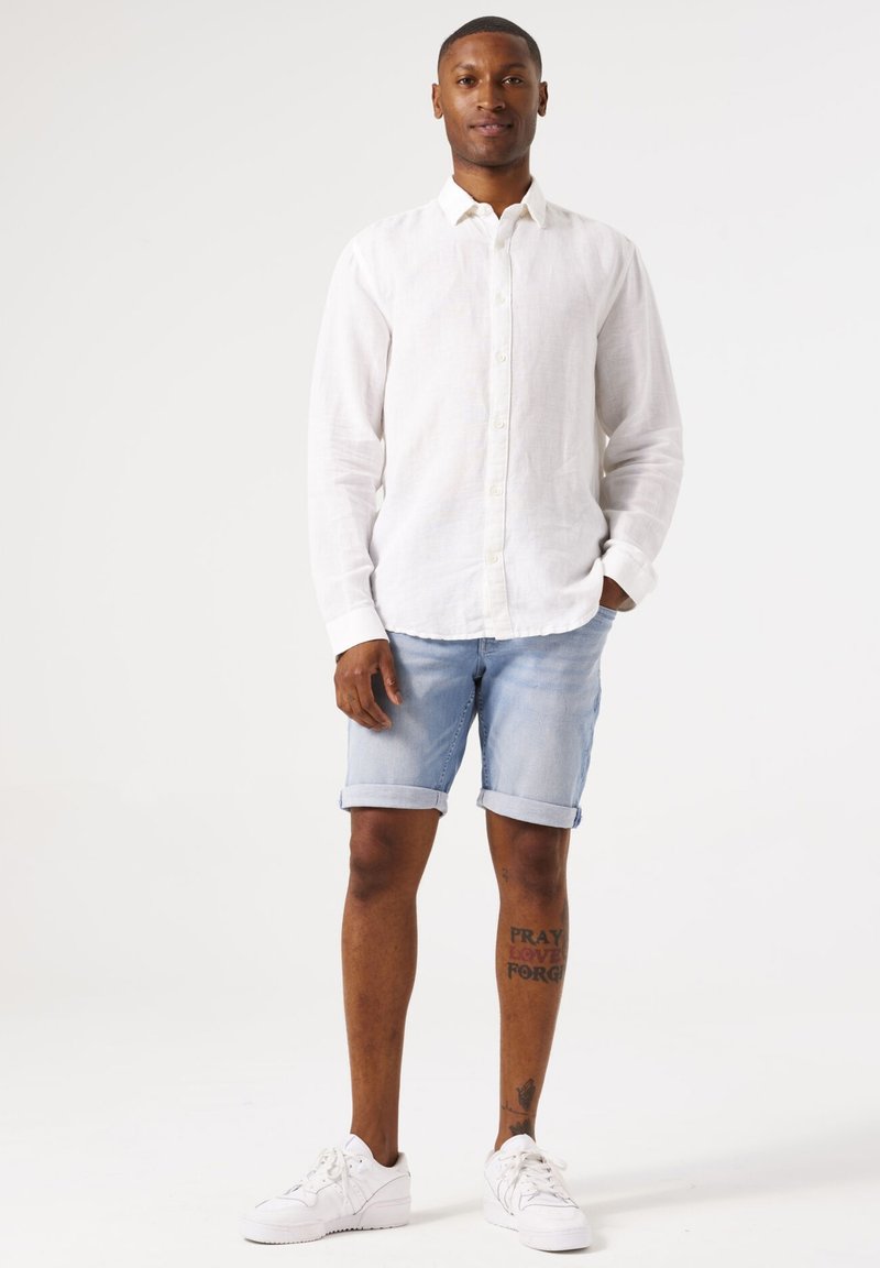 Witte linnen overhemd met knopen, lichtblauwe opgeknoopte denim short, witte sneakers en zichtbare tattoo op het onderbeen. Eenvoudige hedendaagse stijl.