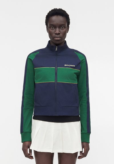 Femme portant une veste zippée bleu marine et verte avec le texte "Palm Angels", associée à une jupe plissée blanche, debout devant un fond uni.