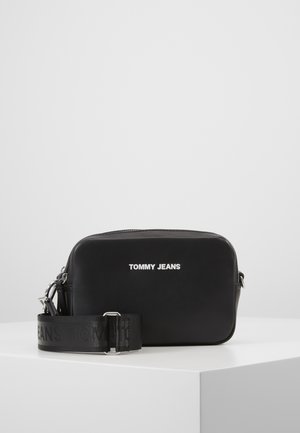 Zwarte Tommy Jeans rechthoekige crossbodytas met brede verstelbare band en zilveren hardware, geplaatst op een witte ondergrond tegen een lichte achtergrond.