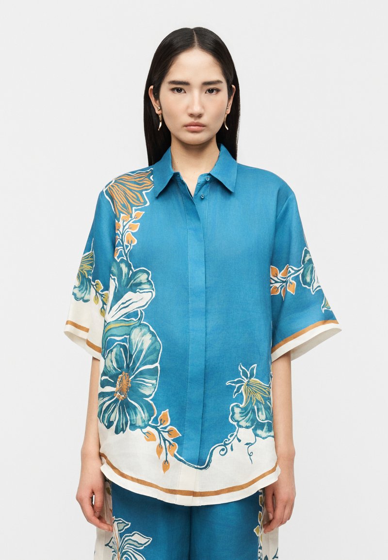 Γυναίκα με μακριά σκούρα μαλλιά, φοράει μπλε υπερμεγέθη πουκάμισο και αντίστοιχο παντελόνι με μεγάλα λουλουδένια σχέδια σε άσπρο, πράσινο και πορτοκαλί.