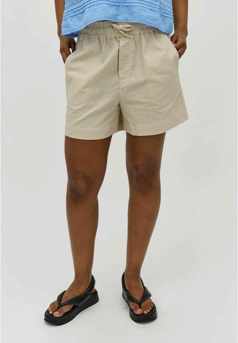 Beige shorts met een elastische tailleband, zijzakken en een relaxte pasvorm, gedragen met zwarte slippers op blote voeten.