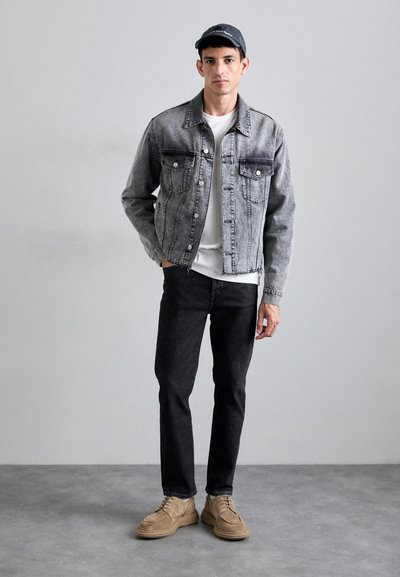Gray denim jacket with two chest pockets, worn over a white shirt, paired with dark jeans and tan suede shoes. Cap completes the outfit.