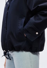 Άτομο που φοράει μια ναυτική μπουφάν με μανσέτες με κουμπιά και ρυθμιζόμενο στρίφωμα, με το χέρι στην τσέπη, συνδυασμένο με ανοιχτόχρωμα μπλε τζιν.