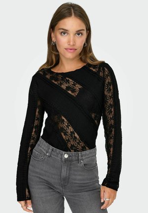 Zwarte longsleeve bodysuit van kant met gestructureerde patronen en diagonale ontwerpelementen, gedragen met grijze high-waisted jeans.