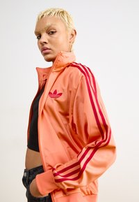 Orangene Satin-Trackjacke mit roten Streifen und Logo, hohem Kragen, Reißverschluss vorne und seitlichen Taschen. Kombiniert mit schwarzem Oberteil und dunkler Jeans.