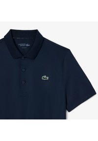 Marinblå polotröja i andningsbart material. Har en klassisk krage, knäppning med tre knappar och Lacoste-logotyp i grönt och rött. Regular fit.