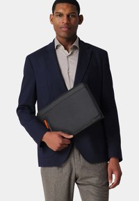 Schwarzes Lederportemonnaie mit orangefarbenem Reißverschlussdetail, rechteckiger Form und glatter Textur, gehalten von einer Person in einem marineblauen Blazer und einem hellen Hemd.