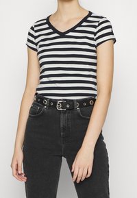 Pruhované tričko s výstřihem do V v námořnické modři a bílé barvě, krátké rukávy, spárované s černými džíny s vysokým pasem a černým opaskem se stříbrnými kovovými detaily.