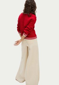 Roter gestrickter Pullover mit einem strukturierten Muster, gerippten Bündchen und Saum; kombiniert mit weiten cremefarbenen Hosen, die einen entspannten, geschichteten Look zeigen.