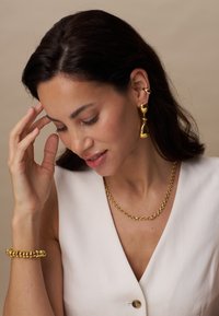Goldene Ohrringe mit Tropfendesign, passende Kettenhalskette und Armband. Glatte Textur, glänzende Oberfläche, getragen mit einem weißen ärmellosen Oberteil.