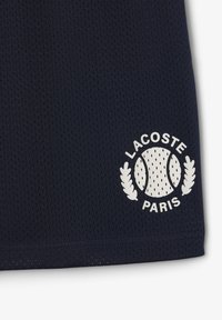 Polo de tejido de malla azul marino con un logo circular blanco que presenta una pelota de tenis, hojas y el texto "LACOSTE PARIS" en la esquina inferior izquierda.
