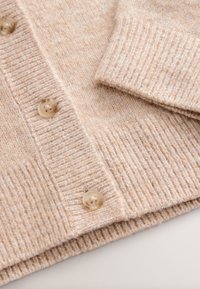 Cardigan en tricot marron clair avec texture côtelée, comportant quatre grands boutons beiges et un détail de bord replié. Tissu doux et chaud.