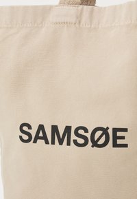 Beige Canvas-Shopper-Tasche mit texturierter Oberfläche, auf der das schwarz gedruckte Logo "SAMSOE" prominent angezeigt wird. Langlebiges, schlichtes Design.