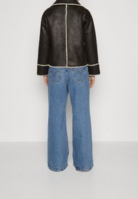 Zwarte leren jas met witte decoratieve stiksels, gecombineerd met lichtblauwe wijde jeans. De achterkant van de outfit is zichtbaar.