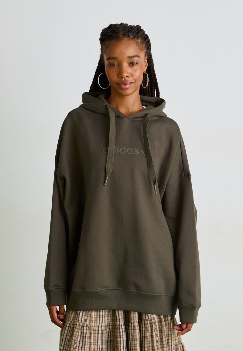 Olivgrön oversized hoodie med dragsnören, utrustad med en subtil präglad logotyp. Mjuk tyg och ribbade ärmslut, tillsammans med en rutigt kjol.