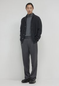 Giovane uomo in piedi contro uno sfondo neutro, indossa un cardigan scuro con bottoni, un dolcevita grigio, pantaloni grigi e scarpe nere, con le mani in tasca.