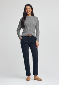 Maglione grigio a maglia con maniche lunghe, abbinato a pantaloni blu navy, una cintura in pelle marrone e mocassini beige, con dettagli decorativi minimi.