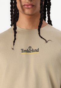 T-shirt en coton beige à manches courtes, avec un logo Timberland noir et jaune centré sur la poitrine. Texture lisse et coupe décontractée.