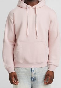 Felpa con cappuccio rosa chiaro in tessuto morbido, con tasca a marsupio, coulisse e polsini e orlo a coste. Indossata sopra jeans blu sbiaditi.