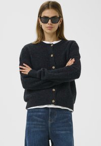 Tmavě modrý pletený cardigan s kulatým výstřihem, osmi knoflíky a dlouhými rukávy. Nositelný přes bílou košili, doplněný modrými džínami.