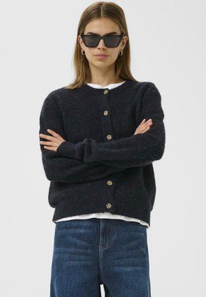 Dunkelblauer Strickcardigan mit rundem Ausschnitt, acht Knöpfen und langen Ärmeln. Getragen über einem weißen Hemd, kombiniert mit blauen Jeans.
