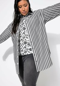 Camicia a righe bianco e nero con maniche lunghe, bottoni e taschino. Indossata sopra un top a fantasia floreale, abbinata a pantaloni grigio scuro.