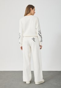 Sudadera de algodón blanca con pantalones a juego, adornados con bordados florales negros en las mangas y el bolsillo trasero, con un corte relajado y un diseño de pierna ancha.