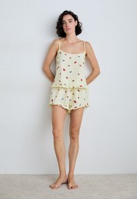 Pajama set s cvjetnim uzorkom u mekoj žutoj tkanini, sa naramenicama, volanastim rubovima i uzorkom trešanja u crvenoj i plavoj boji na gornjem dijelu i kratkim hlačama.