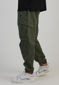 Gröna cargobyxor med flera fickor, elastiska mudd och en subtil logodetalj. Bärs med vita sneakers som har grå accenter.