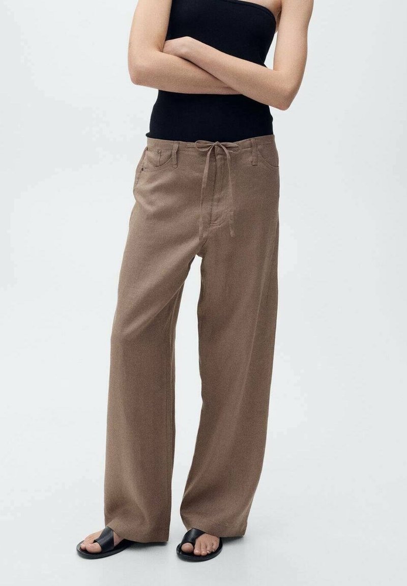 Personne portant un pantalon marron à jambes larges avec taille à cordon, un top noir sans manches et des sandales noires à bouts ouverts, debout, bras croisés.