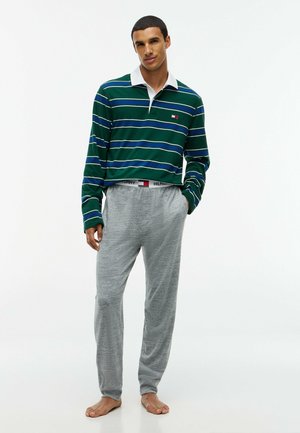 Jonge man staat blootsvoets, draagt een groen gestreept shirt met lange mouwen en grijze joggingbroek, handen in de zakken tegen een witte achtergrond.