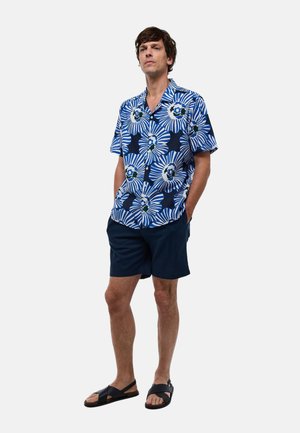 Homme debout, les mains dans les poches, portant une chemise à fleurs bleue à manches courtes, un short bleu marine et des sandales noires sur un fond blanc.