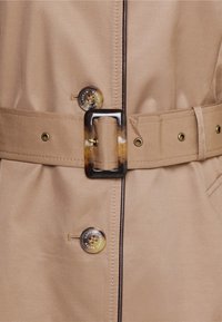 Béžový kabát s páskem a lesklou texturou, který má velké kulaté knoflíky a obdélníkovou sponu se skvrnitým vzorem.