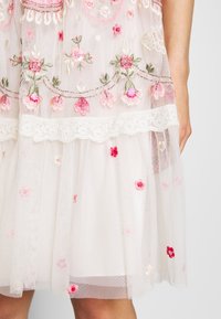 Jupe blanche transparente avec broderies florales roses et rouges, bordure en dentelle, et une main partiellement visible reposant à côté.