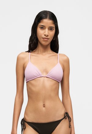 Femme aux longs cheveux foncés portant un haut de bikini rose clair et un bas de bikini noir à nouer sur les côtés, debout devant un fond clair uni.