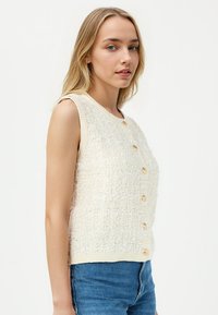 Beige, textuurloze mouwloze top met een ronde halslijn, voorzien van gouden knopen aan de voorkant en een aansluitende zoom. Gemaakt om te dragen over blauwe denim.