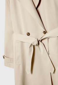Beżowy płaszcz trench z podwójnym zapięciem, dużymi guzikami, paskiem w talii oraz kieszeniami po bokach. Gładka tekstura materiału.