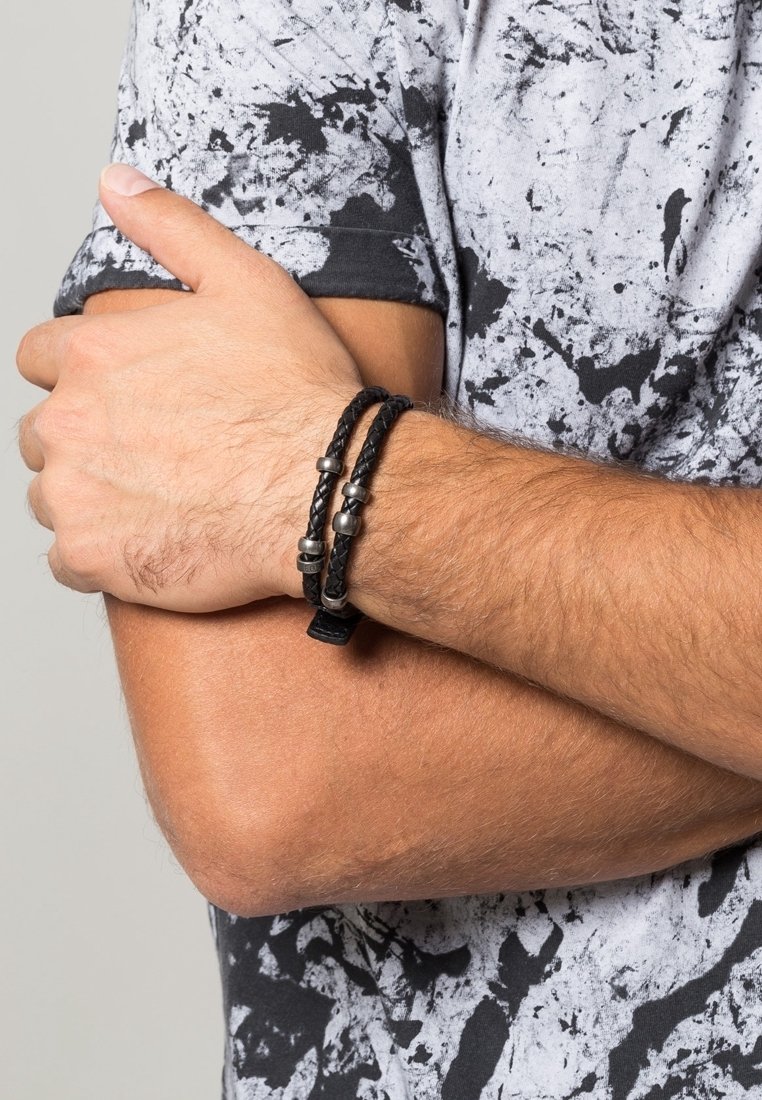 Bracelet double tour en cuir noir tressé avec des perles métalliques, porté au poignet d'une personne croisant les bras sur une chemise éclaboussée de noir et blanc.