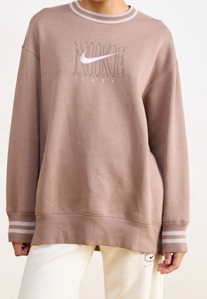 Person iført en løs brun Nike sweatshirt med striped manchetter og hvidt Nike-logo og tekst på brystet, kombineret med lyse bukser.
